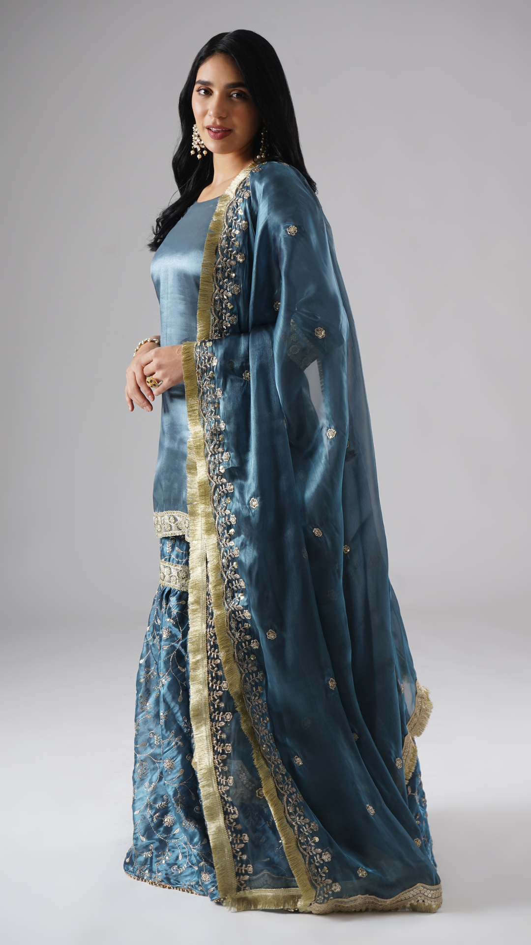Model styled in the Tehzeeb-e-Aab teal blue gharara set by Zahrat, side view highlighting the flowing dupatta and full outfit.