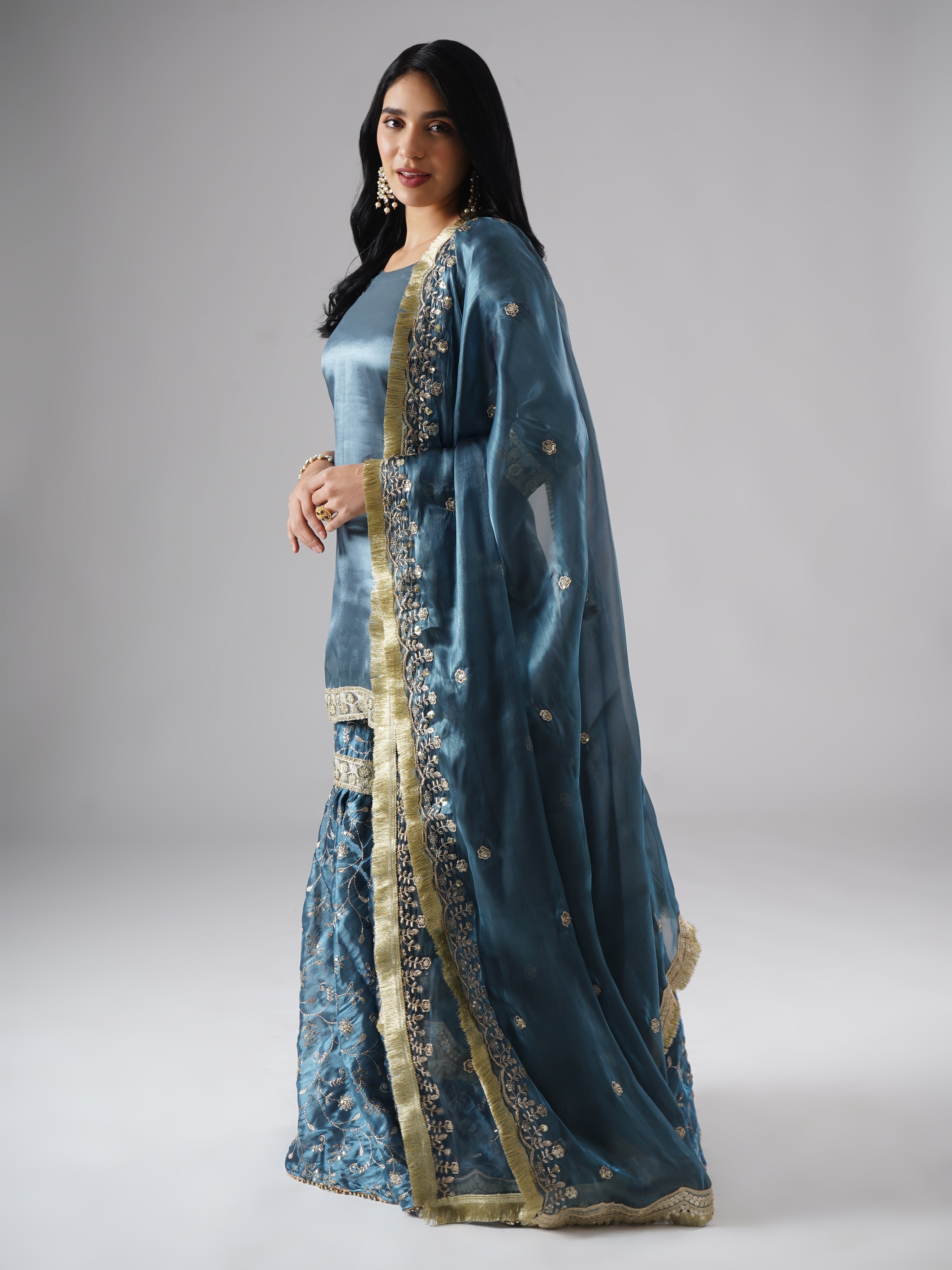 Model styled in the Tehzeeb-e-Aab teal blue gharara set by Zahrat, side view highlighting the flowing dupatta and full outfit.