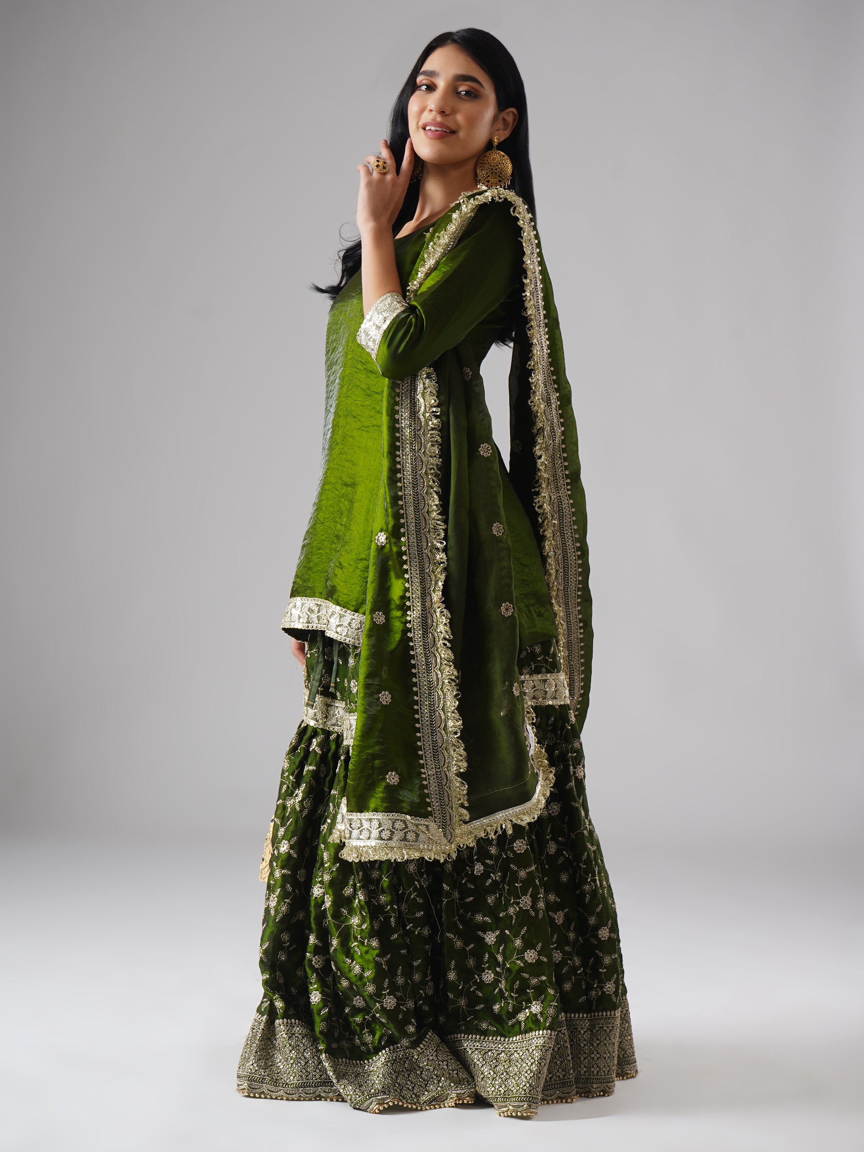 Model styled in the Khushrang Henna Gharara by Zahrat, side view highlighting a festive dupatta drape and the full outfit in henna green.