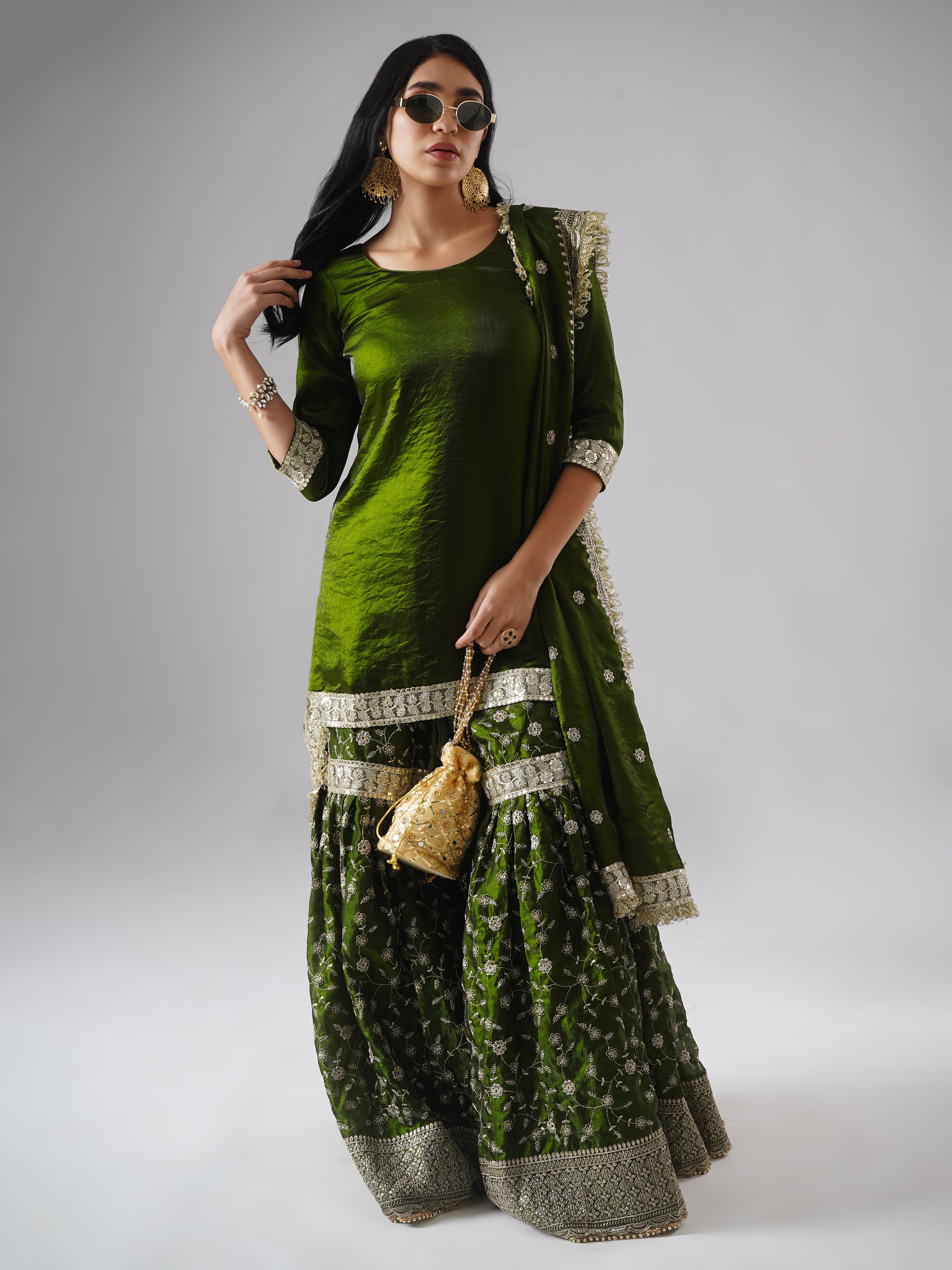 Lifestyle shot of model wearing Zahrat’s Khushrang Henna jewel-toned henna green gharara set, styled with a potli bag and sunglasses.