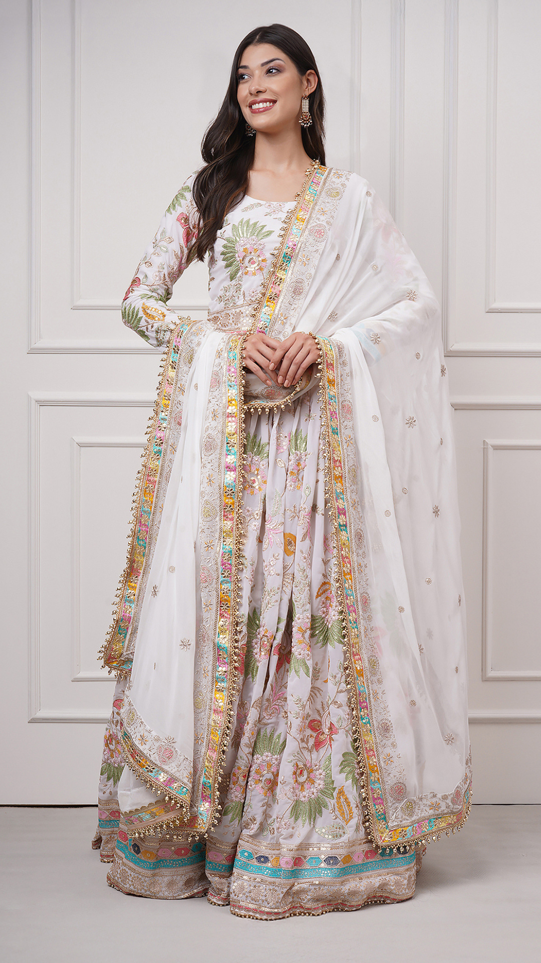 A woman models Eira Bloom Lehenga Set - Zahrat Lehenga with a floral print and ornate multicolor borders, standing against a white paneled wall. Front view