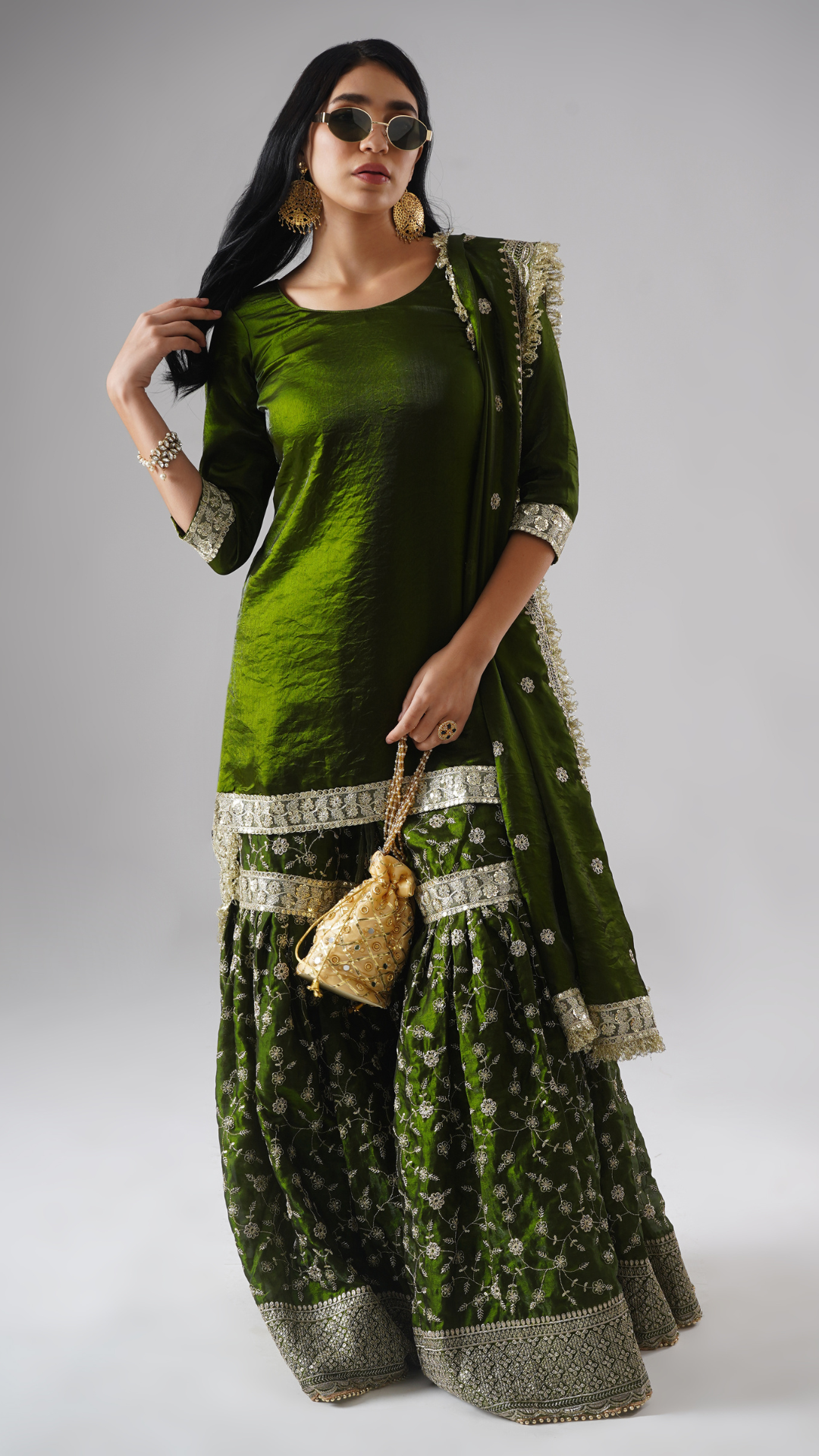 Lifestyle shot of model wearing Zahrat’s Khushrang Henna jewel-toned henna green gharara set, styled with a potli bag and sunglasses.