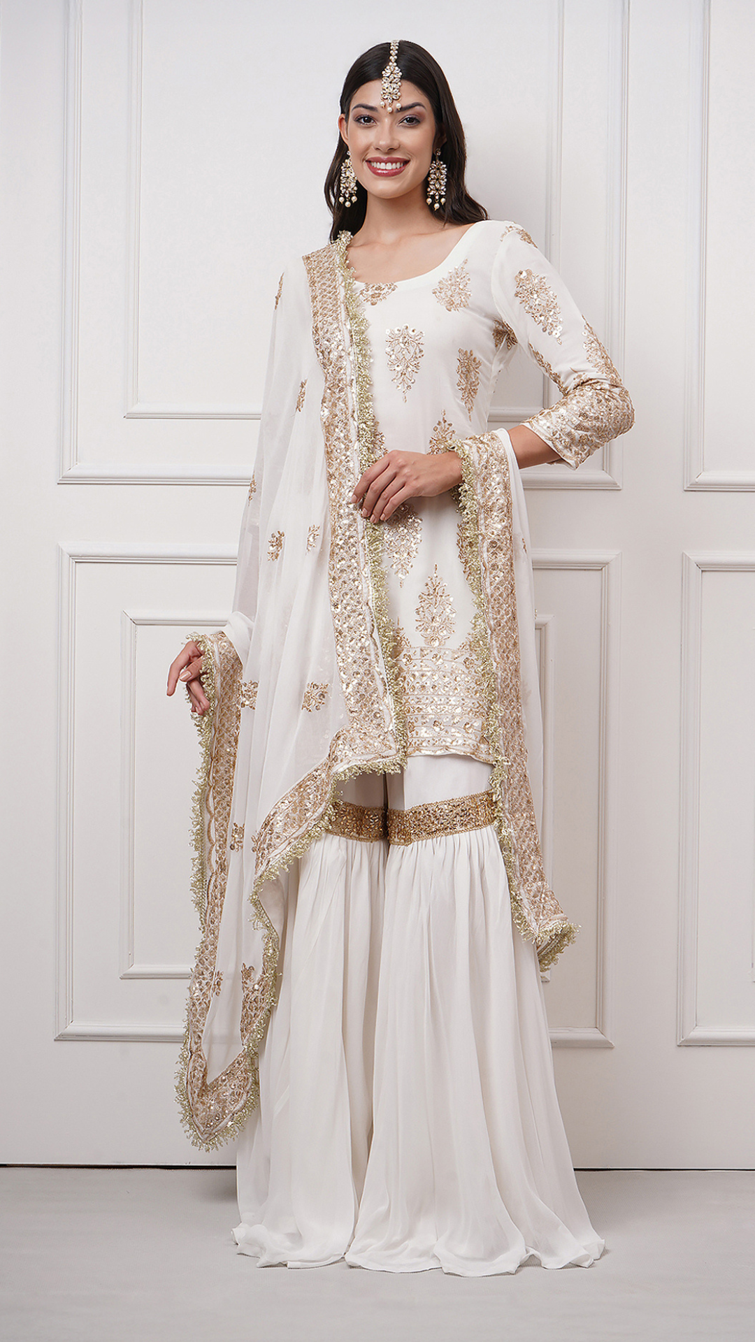 A woman models in Naura – White Embroidered Gharara Set by Zahrat showcasing the gold embroidered kurta, dupatta and a plain white flared harara.