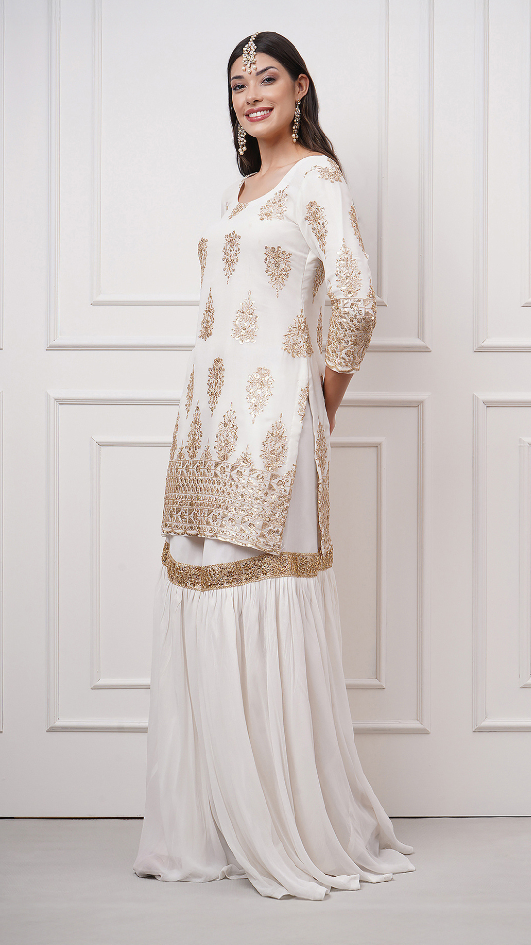 A woman models in Naura – White Embroidered Gharara Set by Zahrat showcasing the gold embroidered kurta and a plain white flared gharara without dupatta.