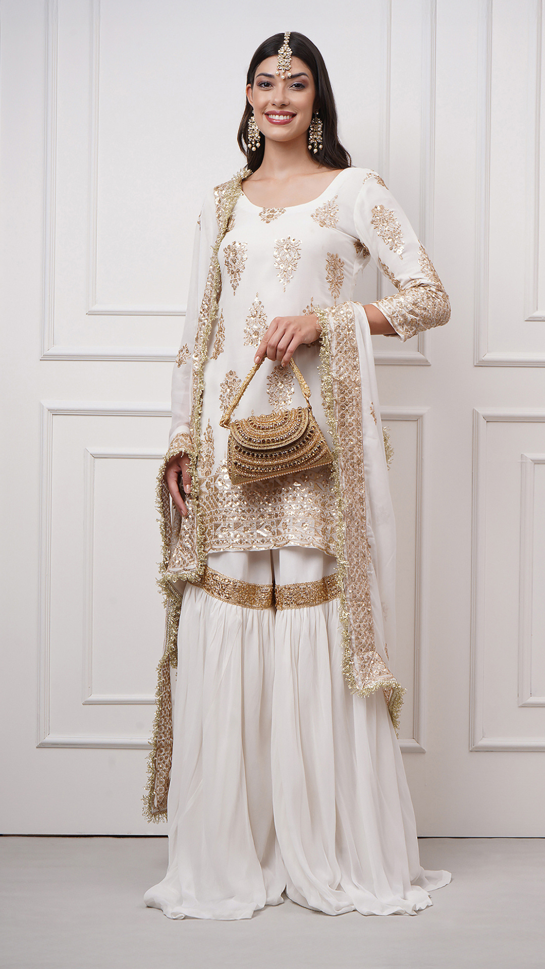 Lifestyle photo of a model wearing the Naura – White Embroidered Gharara Set by Zahrat showcasing the gold embroidered kurta, dupatta and a plain white flared gharara.