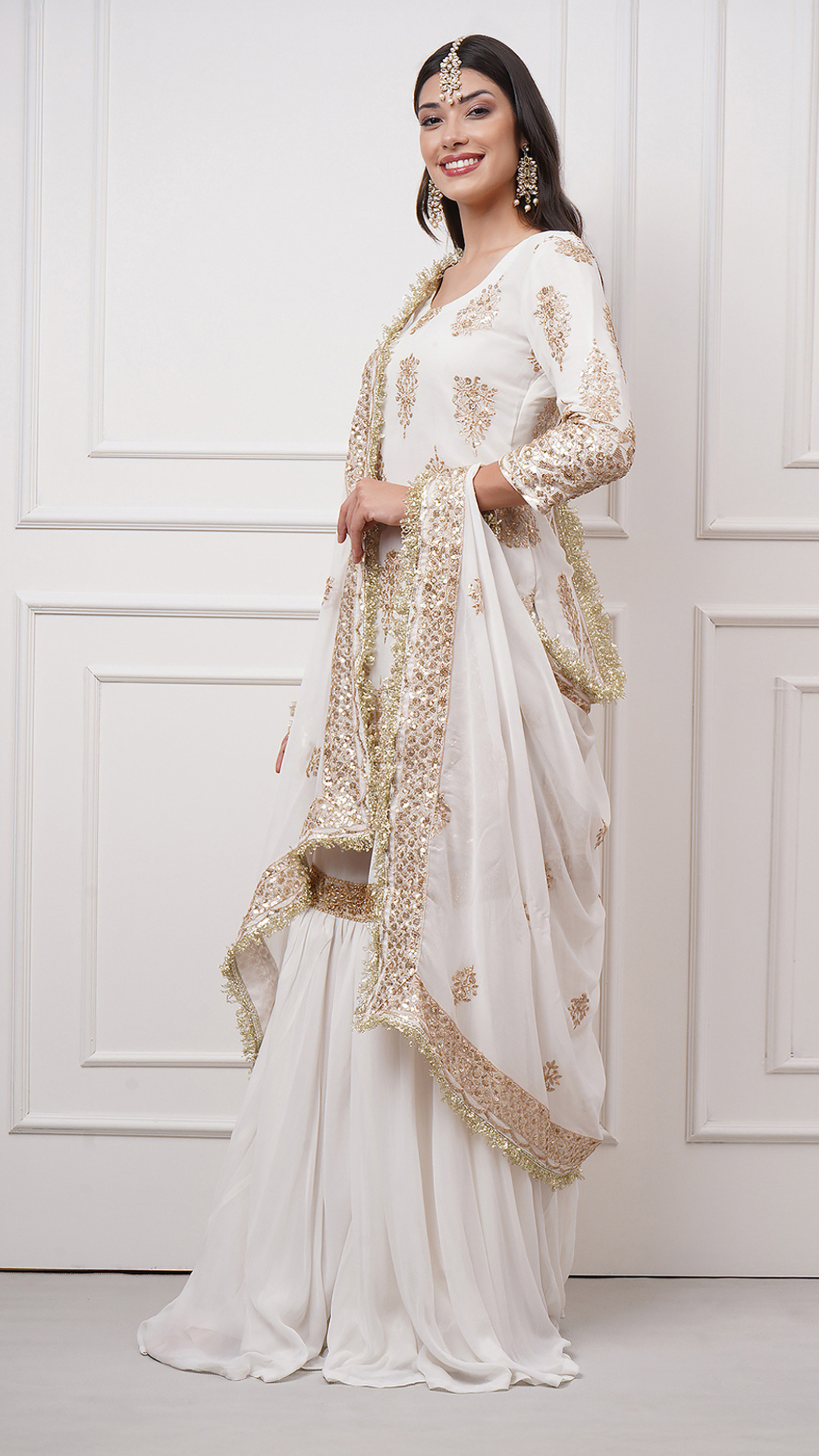 A woman models in Naura – White Embroidered Gharara Set by Zahrat showcasing the gold embroidered kurta, dupatta and a plain white flared harara. - Left side view.