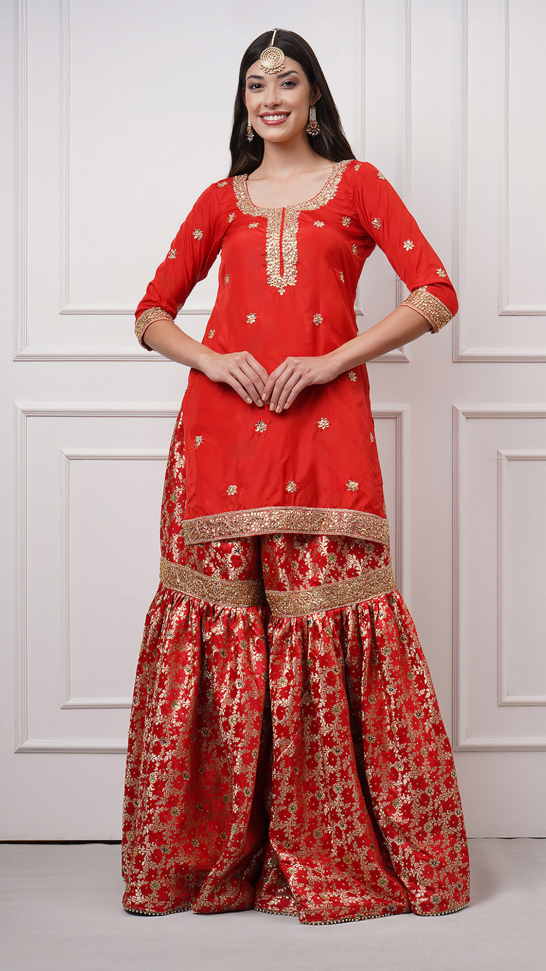 A women models in Surkhnaaz – Red Kimkhwab Bridal Gharara Set by Zahrat showcasing the stone embellished kurta and gharara. 