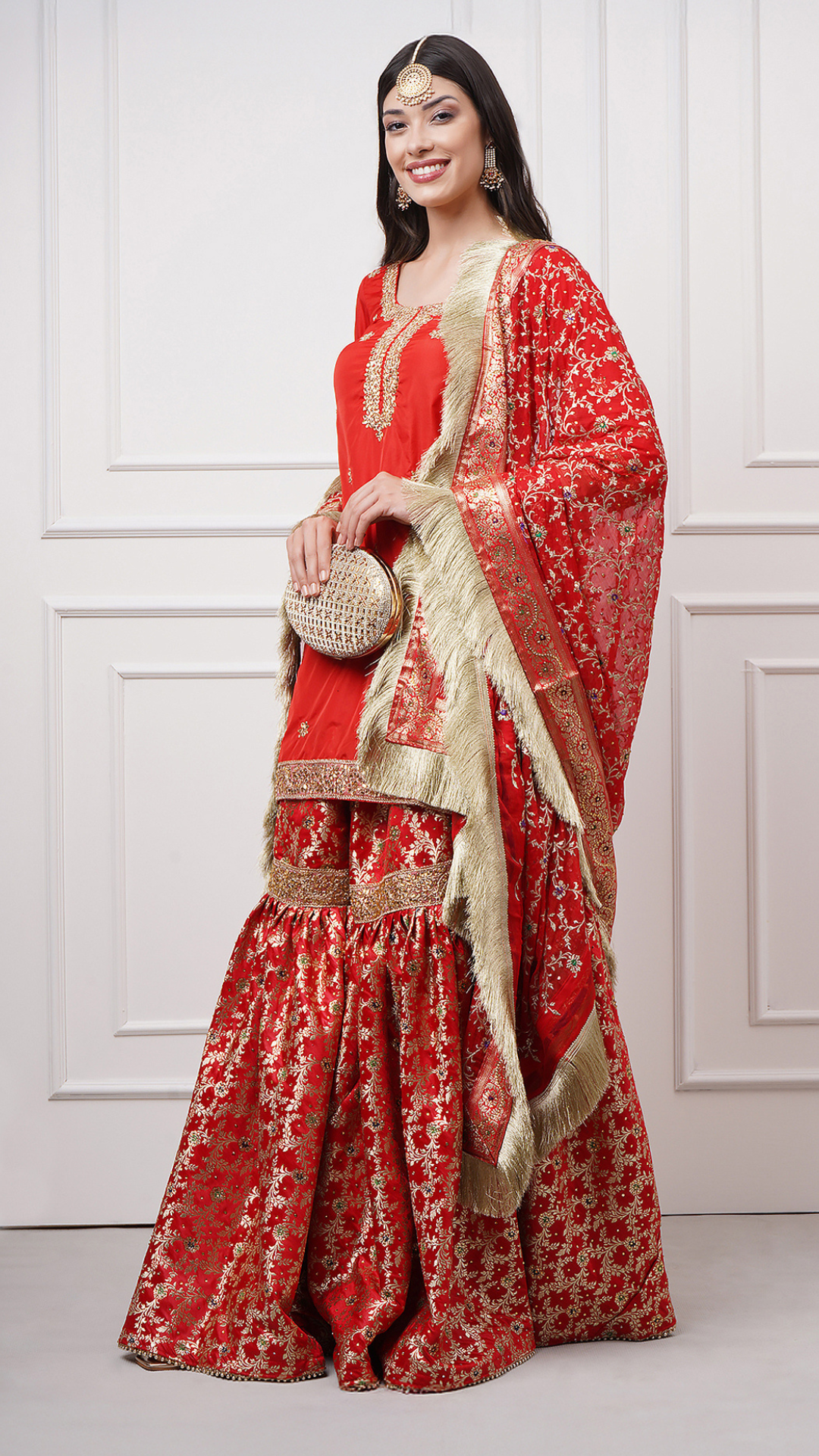 A women models in Surkhnaaz – Red Kimkhwab Bridal Gharara Set by Zahrat. A lifestyle shot with the embroidered potli bag.