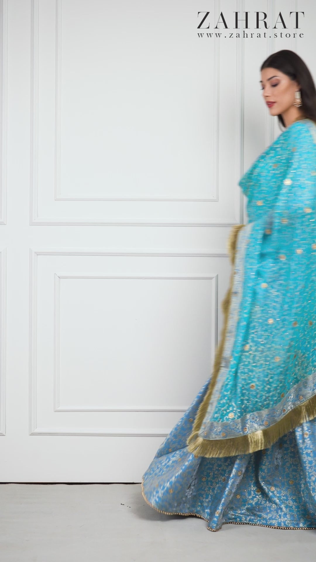 Video of a model wearing the Zahrat Falaknuma – Sky Blue Kimkhwab Gharara Set, walking gracefully and twirling to display the gharara’s wide flare. The golden zari and stone embellishments catch the light, and the sheer dupatta glides with movement, revealing shimmering lace borders and gold fringe in motion. 
Zahrat